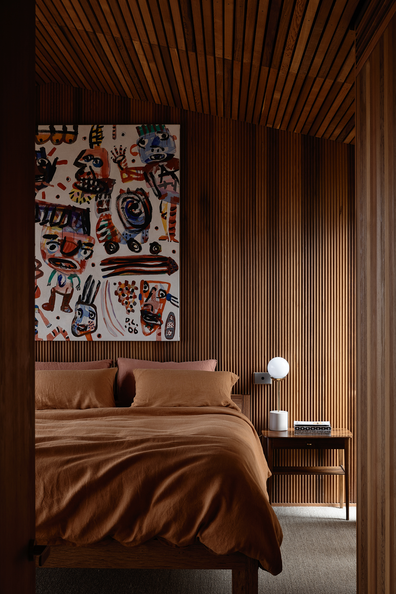 A bedside table, bed and painting in a bedroom with timber walls.