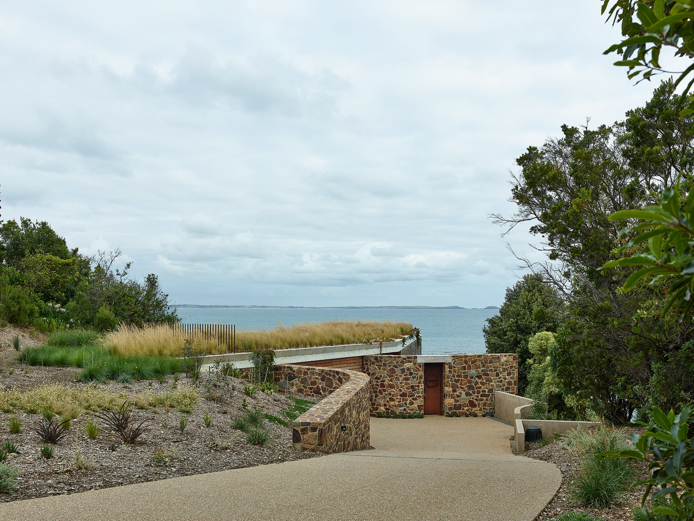 A stone walkway leading to a building with a grass roof. There is a body of water in the background.