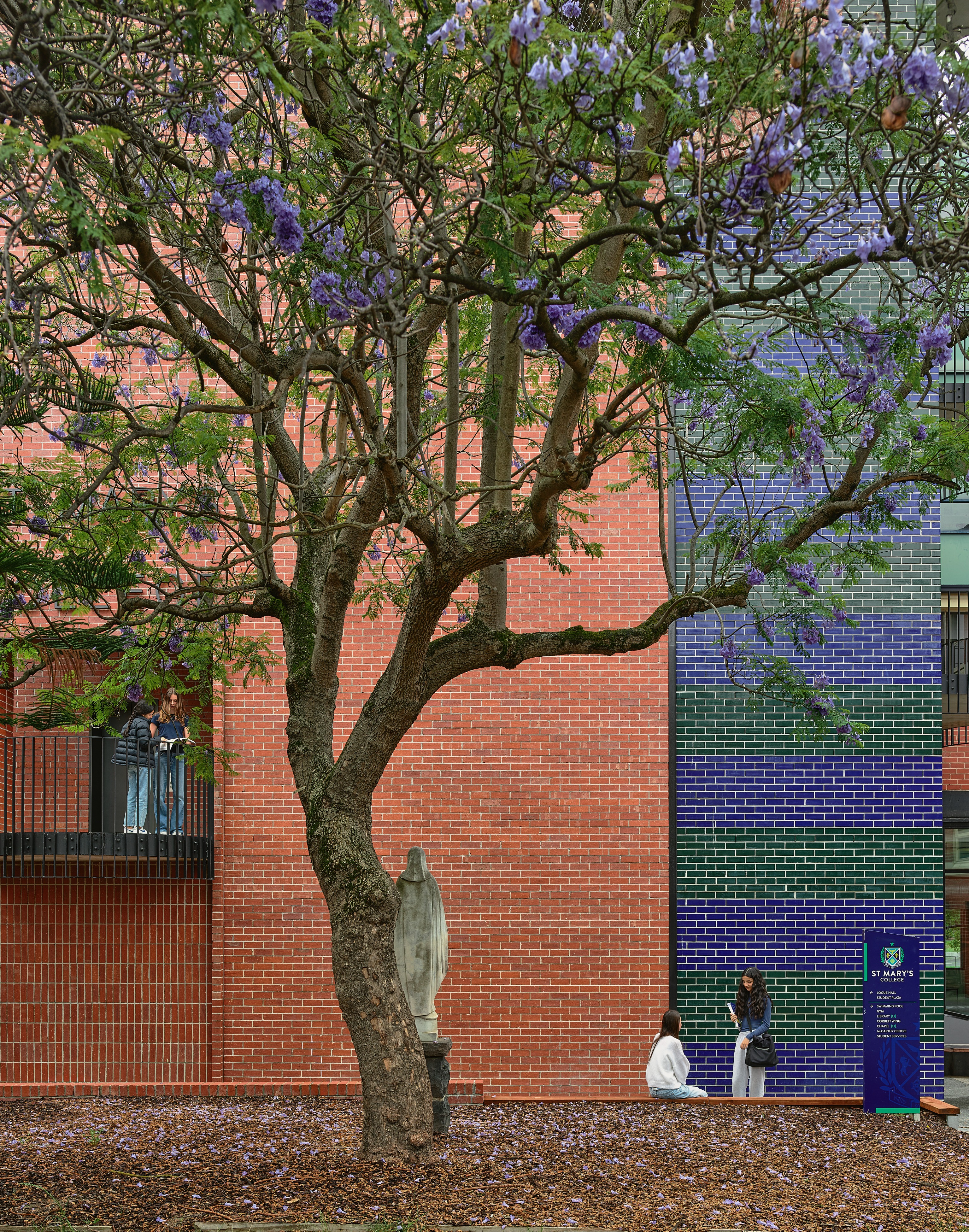 A tree in the foreground, with a red brick building and people chatting in the background.